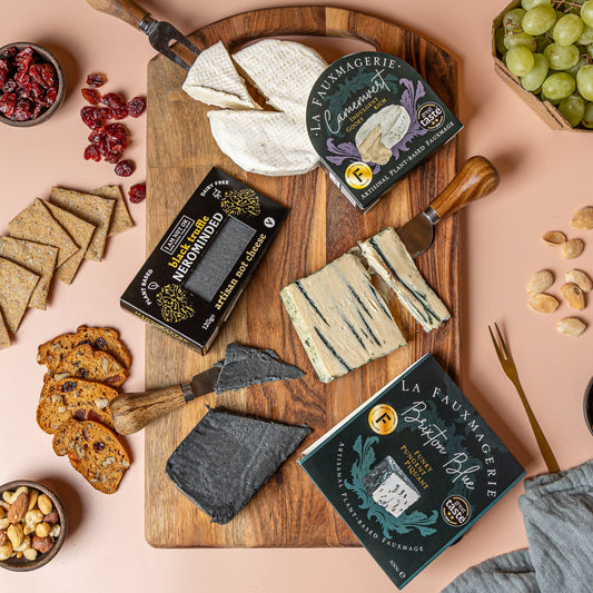 Assorted cheeses on a wooden board with packaging and accompaniments on a pink background. Camemvert and Brixton Blue by La Fauxmagerie and the Nerominded by I am nut ok.